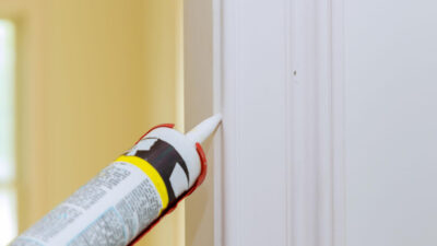 Close-up of a small gap where a painted door casing meets an interior wall, with a caulk gun and sealant ready for repair.