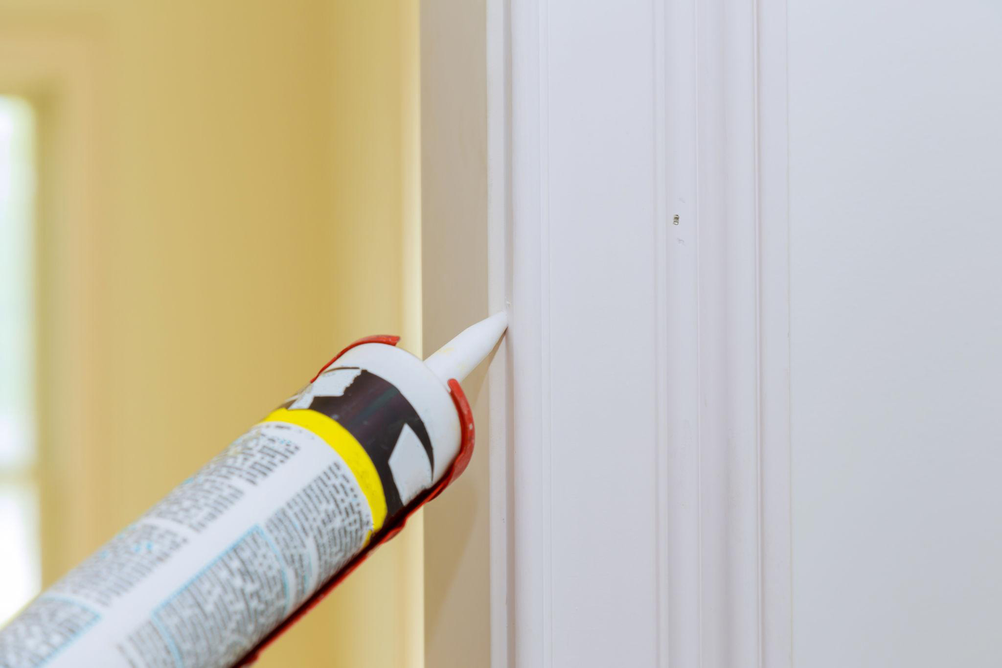 Close-up of a small gap where a painted door casing meets an interior wall, with a caulk gun and sealant ready for repair.